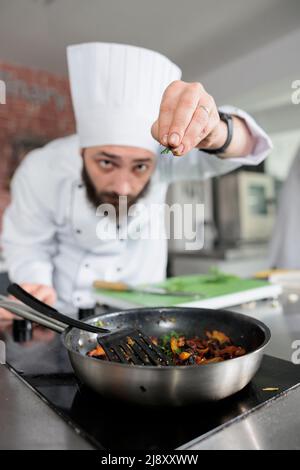 Un expert gastronomique jette des herbes fraîches hachées dans une casserole tout en cuisinant un plat gastronomique pour le service du dîner au restaurant. Chef cuisinier préparant le repas du soir dans une cuisine professionnelle avec des légumes biologiques. Banque D'Images