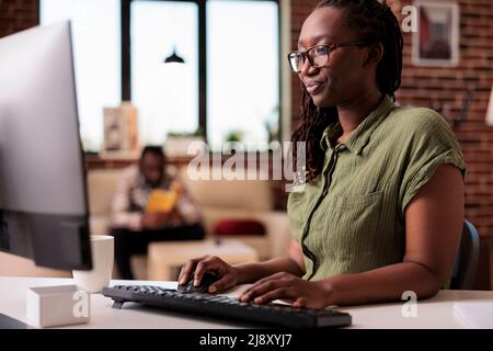Un programmeur afro-américain spécialisé travaillant à distance de chez lui pendant que le petit ami lit un livre sur un canapé. Indépendant regardant l'écran d'ordinateur et la dactylographie en utilisant pc dans le salon de maison. Banque D'Images