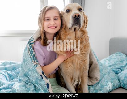 Petite fille avec chien Golden Retriever enveloppée dans une couverture dans le lit dans la chambre. Jolie petite fille qui s'embrasse chien chien labrador à la maison Banque D'Images