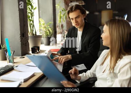 Processus de travail au sein d'une entreprise moderne. Un jeune homme et une jeune femme discutent de nouvelles tâches assis à une table de bureau, à l'intérieur. Concept d'équipe, de poste, de carrière Banque D'Images