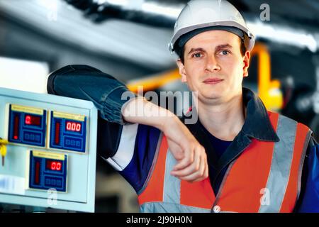Portrait d'un jeune ingénieur de l'industrie lourde dans un gilet de protection, des vêtements de travail et un casque. Un travailleur confiant regarde directement dans la caméra. Usine technologique moderne ou usine de production. Arrière-plan Banque D'Images