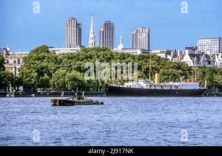 1982 image d'archive des gratte-ciel de Londres vue à travers la Tamise depuis South Bank - montrant les tours du Barbican, la flèche de l'église de St Bride et le dôme de l'Old Bailey. Le bateau amarré sur la rive nord est le HMS Chrysanthemum, une boucle de classe Anchusa de 1917, mis au rebut en 1995. Banque D'Images