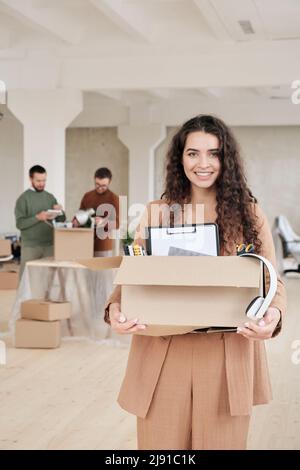 Portrait d'une jeune femme d'affaires souriante aux cheveux bouclés dans un élégant coffret de costume avec bloc-notes, presse-papiers et écouteurs tout en se déplaçant dans un nouveau bureau Banque D'Images
