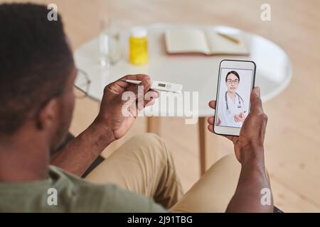 Homme afro-américain méconnaissable contrôlant la température corporelle tout en consultant son médecin en ligne à l'aide d'un smartphone Banque D'Images