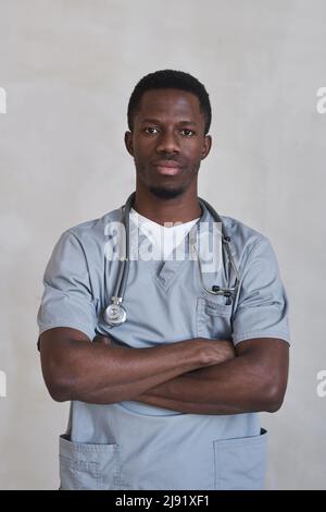 Portrait vertical moyen d'un beau médecin noir portant un uniforme gris debout avec les bras croisés regardant la caméra Banque D'Images