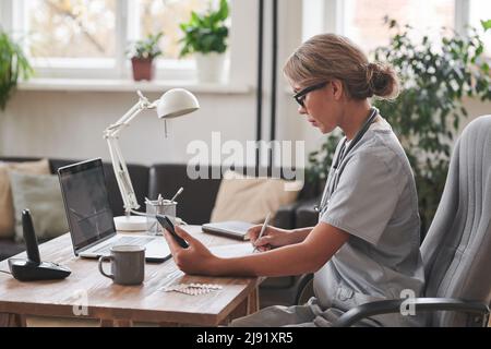 Vue latérale moyenne prise de vue d'une femme thérapeute jeune adulte assise sur un bureau devant un ordinateur portable tenant un smartphone pour prendre des notes Banque D'Images