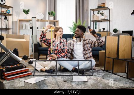 Heureux couple multiculturel assis au sol dans le salon, regardant la caméra et tenant divers outils pour assembler des meubles. Concept de jeune famille se déplaçant dans une nouvelle maison. Banque D'Images