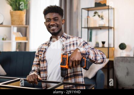 Portrait d'un jeune homme afro-américain qui assemble des meubles à la maison à l'aide d'un tournevis sans fil. Homme souriant s'amusant en rénotant son nouvel appartement. Banque D'Images