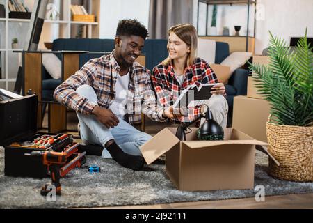 Jeune famille multiraciale assise dans la salle de séjour et déballant les boîtes en carton ensemble. Bonne épouse caucasienne et mari afro-américain souriant sincèrement tout en tenant le cadre photo. Banque D'Images