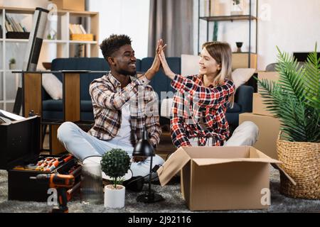 Beau homme afro-américain et belle femme caucasienne faisant cinq grands gestes tout en étant assis sur le sol dans le salon avec des boîtes autour. Concept de famille et de réinstallation. Banque D'Images