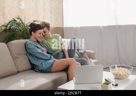 les amateurs de pansexués heureux assis sur un canapé près d'une table basse avec un ordinateur portable et du pop-corn Banque D'Images