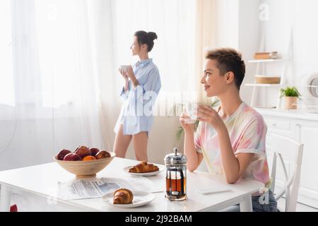personne pansexuée souriante assise avec une tasse de thé près des croissants et partenaire sur fond flou Banque D'Images