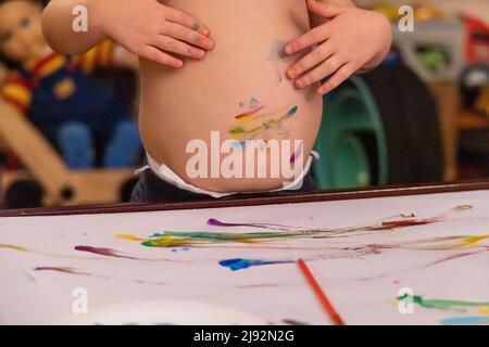 Drôle de garçon de deux ans, est vu sans une chemise essuyant la peinture des mains couvertes sur l'estomac. Un enfant en désordre aime peindre avec de la place pour la copie. Banque D'Images