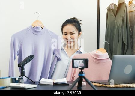 De jeunes entrepreneurs asiatiques ramassant leurs vêtements pour les montrer à leurs clients par le biais de ventes en ligne sur un téléphone en direct. Banque D'Images