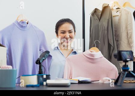 De jeunes entrepreneurs asiatiques ramassant leurs vêtements pour les montrer à leurs clients par le biais de ventes en ligne sur un téléphone en direct. Banque D'Images