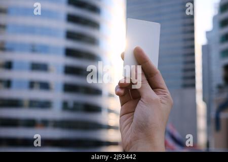 Une main tenant une carte de crédit blanche vierge avec lumière du soleil qui brille à travers sur le côté et les bâtiments élevés en arrière-plan. Banque D'Images