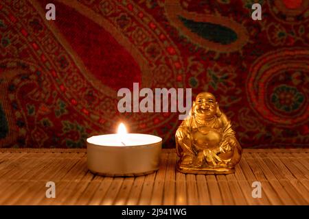 Statue de bouddha de moine doré sur tapis en bois avec fond floral coloré et bougie allumée, gros plan. Concept méditatif Banque D'Images