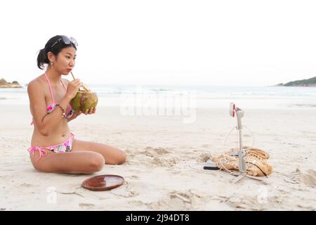 Influenceur buvant un jus de noix de coco tout en faisant un live en utilisant un mobile sur la plage Banque D'Images