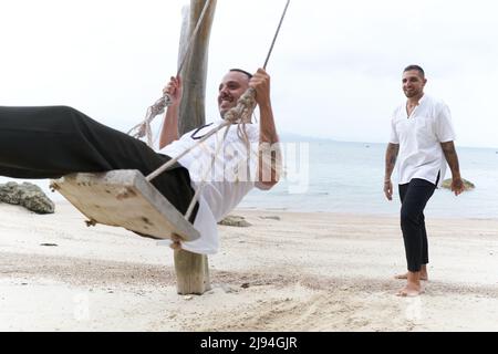 Homme gay balançant sur une balançoire de plage à côté de son couple Banque D'Images