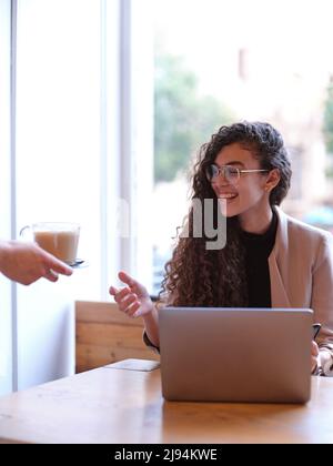 femme souriante recevant un café du serveur tout en travaillant sur son ordinateur, arrière-plan vertical Banque D'Images