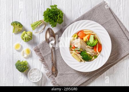 soupe de printemps au poulet avec brocoli, carottes, panais, poireaux et pâtes dans un bol blanc sur une table en bois blanc avec cuillère et ingrédients, vue horizontale fr Banque D'Images