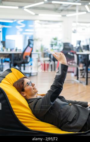 Une femme d'affaires en costume se repose dans un confortable sac de chaise et prend un selfie. Une employée féminine utilise un smartphone et se détend Banque D'Images