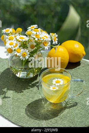 Tisane dans une tasse de verre avec camomille et citrons sur un napperon vert. Thé au citron camomille. Fleurs sauvages en pâquerette dans un pot en verre Banque D'Images