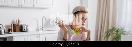 femme tatouée avec la coiffure tendance stylo et pomme fraîche dans la cuisine floue, bannière Banque D'Images