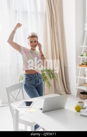 femme excitée montrant le geste de triomphe tout en parlant sur un smartphone près d'un ordinateur portable dans la cuisine Banque D'Images