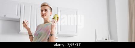 femme souriante avec coiffure tendance tenant pomme et stylo dans la cuisine, bannière Banque D'Images