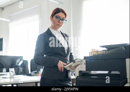 Femme d'affaires en costume et lunettes fait des copies de documents sur une photocopieuse. Une responsable de bureau travaille sur papier. Secrétaire fait Banque D'Images