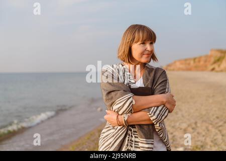 Femme européenne senior marchant avec un livre sur une plage de sable Banque D'Images