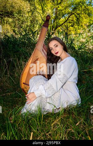 Jeune femme ukrainienne dans une robe blanche et avec les cheveux coulant s'assoit sur l'herbe verte et tient une bandura. Banque D'Images