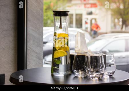 Une carafe en verre transparent, un pichet de limonade avec des citrons, de la menthe à côté des verres sur une table ronde noire dans un café ou un restaurant contre une fenêtre panoramique. A r Banque D'Images