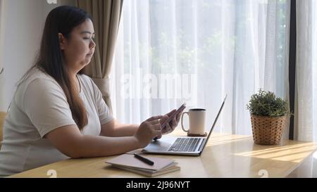 Vue latérale une femme asiatique assise devant un ordinateur portable et utilisant un téléphone portable à la maison Banque D'Images