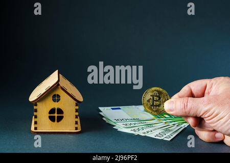 Un homme met une pièce en bitcoin sur une centaine de billets en euros devant une maison en bois sur fond gris Banque D'Images