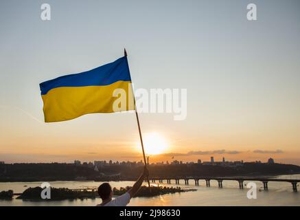 Le grand drapeau ukrainien flotte sur fond de ciel et de paysage urbain au coucher du soleil. Un homme se tient sur le toit de la maison à Kiev et porte le drapeau. Symbo national Banque D'Images