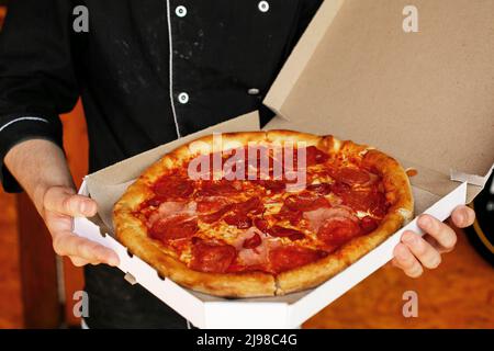 le chef de la pizza porte un tablier noir qui contient de la pizza fraîchement cuite dans une boîte ouverte Banque D'Images
