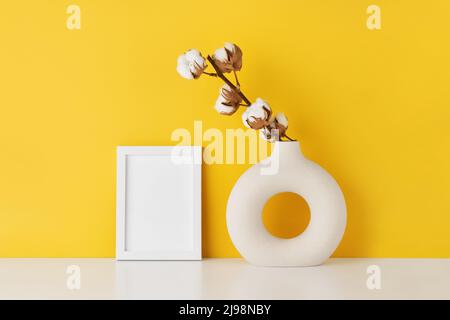 Maquette blanche et vase rond en céramique avec branche de coton sur une table blanche près du mur jaune. Copier l'espace pour le texte Banque D'Images