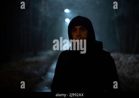 Gars dans le parc la nuit. Homme dans la cagoule noire. Inconnu sur la route la nuit. Portrait dans l'obscurité. Banque D'Images