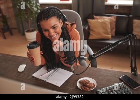 Vue en grand angle d'une jeune femme gaie portant un casque assis au bureau, buvant un latte et lisant des commentaires amusants de ses adeptes Banque D'Images