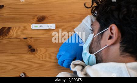 Vue de dessus de l'homme caucasien triste fait un appareil rapide de test à domicile pour Covid19 virus avec un résultat positif et tenant sa tête à la main à la table en bois. Anti Banque D'Images