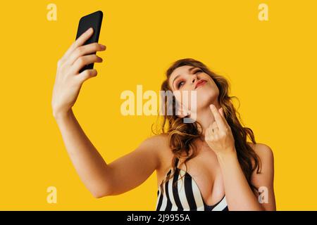 portrait de belle jeune femme sms flirt selfie photo message secret argent signe doigts avec smartphone sur fond jaune studio Banque D'Images