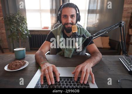 Portrait horizontal d'un jeune vlogger moyen-oriental moderne barbu portant des vêtements décontractés assis au bureau seul dans la chambre loft faisant du contenu looki Banque D'Images