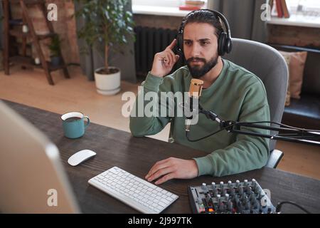 Portrait moyen à grand angle d'un jeune blogueur du Moyen-Orient élégant portant un casque assis sur un bureau parlant sur un appareil photo d'ordinateur Banque D'Images