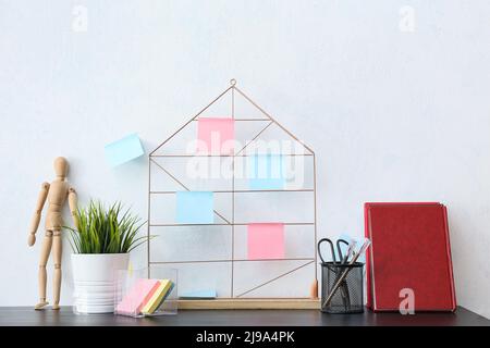 Tableau d'humeur avec différentes notes adhésives, fournitures de papeterie, carnets, poupée en bois et maison sur table sombre près du mur blanc Banque D'Images