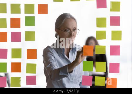 Une femme d'affaires d'âge moyen concentrée prend des notes sur les papiers adhésifs Banque D'Images