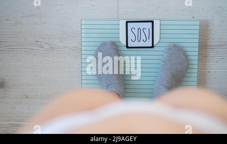 La femme grasse est pesée. Une vue de dessus des pieds femelles en chaussettes grises se trouve sur une échelle électronique. Inscription SOS sur l'affichage de l'échelle de sol. Banque D'Images