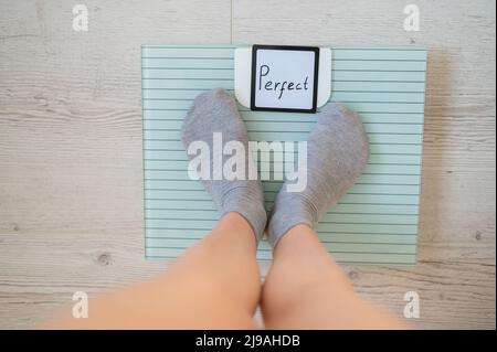 Une femme sans visage mesure le poids sur les balances de sol. Vue de dessus des pieds pour femmes en chaussettes grises sur la balance avec le mot Perfect. L'inscription sur le Banque D'Images