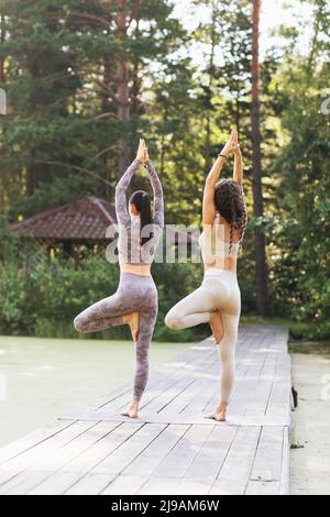 Deux femmes effectuent une variation de l'exercice vrikshasana, posture de l'arbre avec les bras levés, debout sur un pont en bois dans un parc Banque D'Images